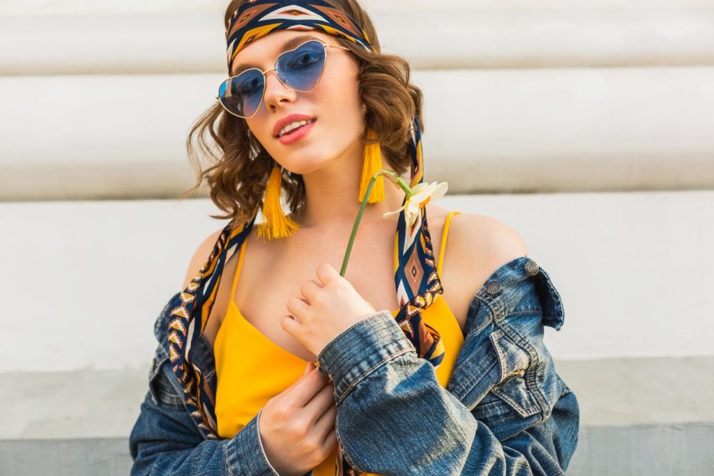 Beautiful and stylish woman in yellow dress and denim jacket posing with blue sunglasses, epitomizing spring and summer fashion trends in street setting, adorned with trendy accessories